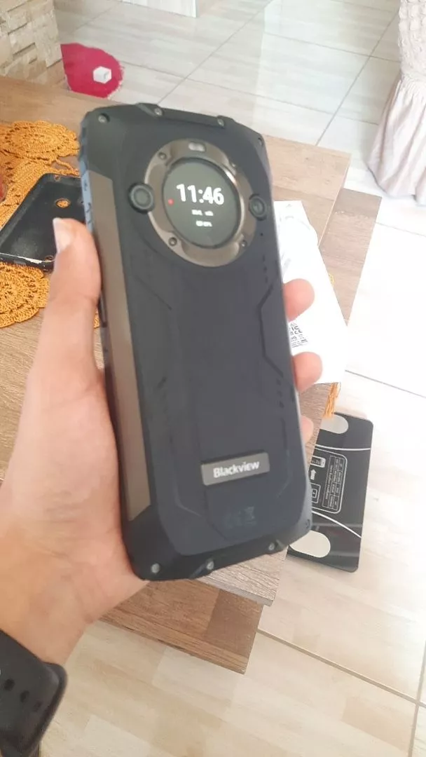 Uma mão segura o smartphone robusto Blackview BV9300 Pro, mostrando a parte traseira preta e texturizada. O destaque é a tela secundária redonda que exibe o horário "11:46". Abaixo da tela secundária, o logo "Blackview" está visível. O fundo é uma sala desfocada com uma mesa de madeira.