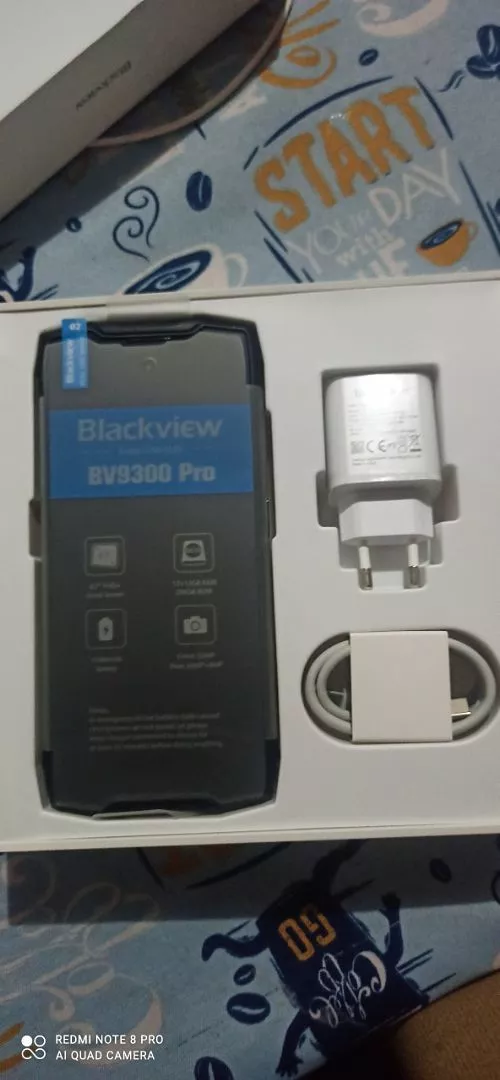 Uma caixa branca aberta mostrando o unboxing do smartphone robusto Blackview BV9300 Pro. O aparelho está deitado à esquerda com uma película protetora de tela azul destacando suas especificações. À direita, estão o adaptador de tomada branco e o cabo USB-C organizados nos compartimentos. A caixa está sobre uma toalha de mesa azul estampada.