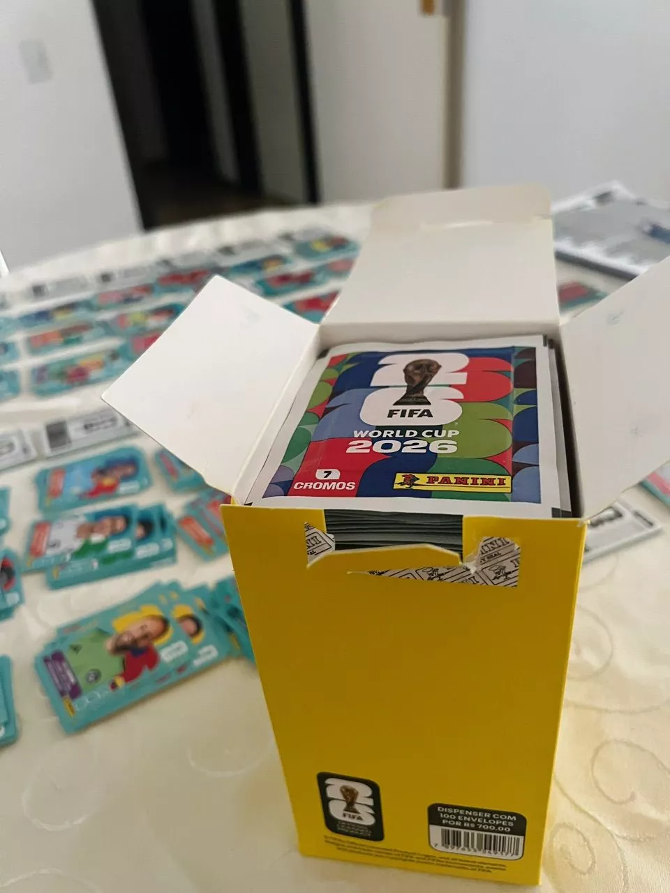 Caixa aberta preta com figurinhas visíveis e rótulo '7 CROMOS', indicando o número de cartas por pacote.