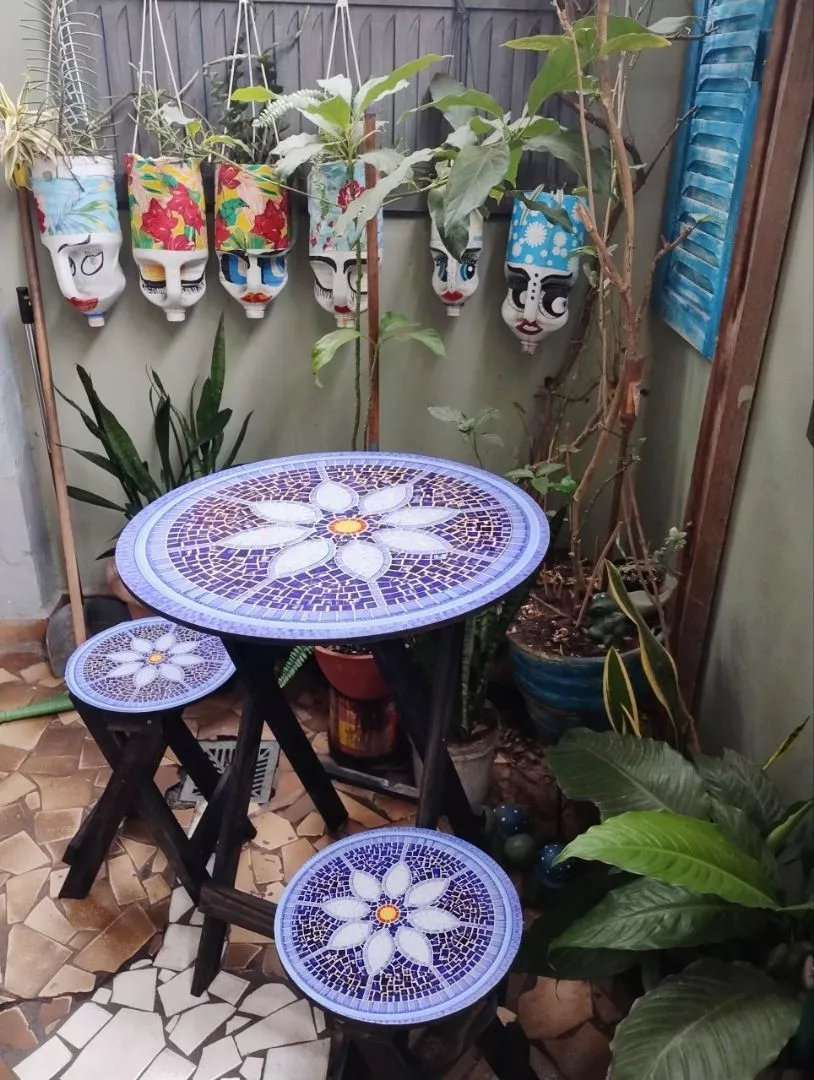 Conjunto de mesa redonda dobrável com 2 bancos em madeira escura, com tampo decorado em mosaico azul e flor branca, em uma área externa com plantas e decorações penduradas.