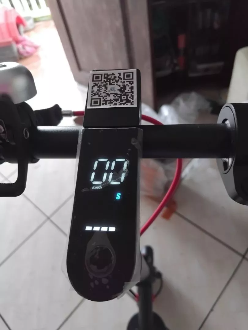 Close-up do display digital do patinete elétrico exibindo "00 km/h" e indicador de bateria, com um adesivo de QR code na parte superior do guidão