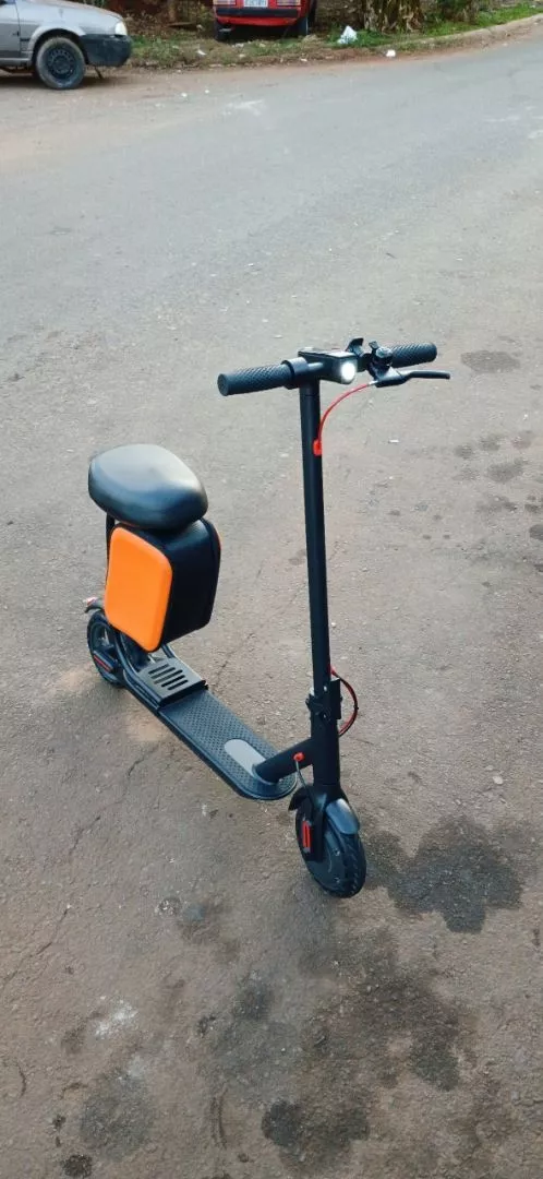 Patinete elétrico preto com banco e bag laranja, estacionado sobre uma estrada de asfalto cinza, com carros e vegetação desfocados ao fundo