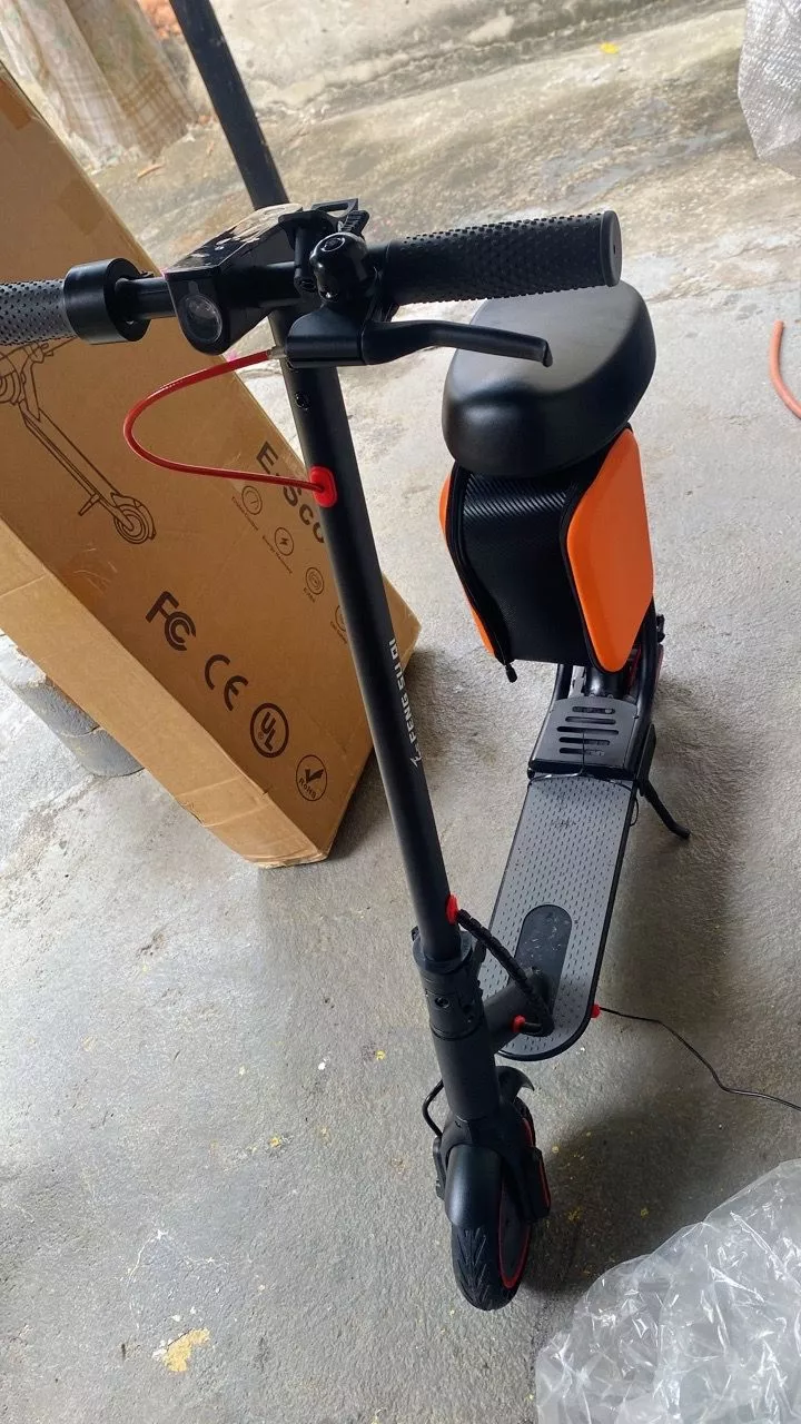 Patinete elétrico preto com banco e bag laranja, parcialmente desembalado, posicionado em um piso de cimento próximo a uma caixa de papelão em ambiente interno
