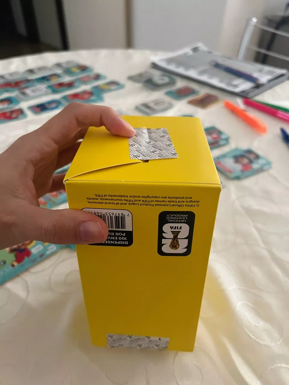 Pacote amarelo com logotipo da FIFA, código de barras e texto '100 envelopes', sendo segurado por uma mão.