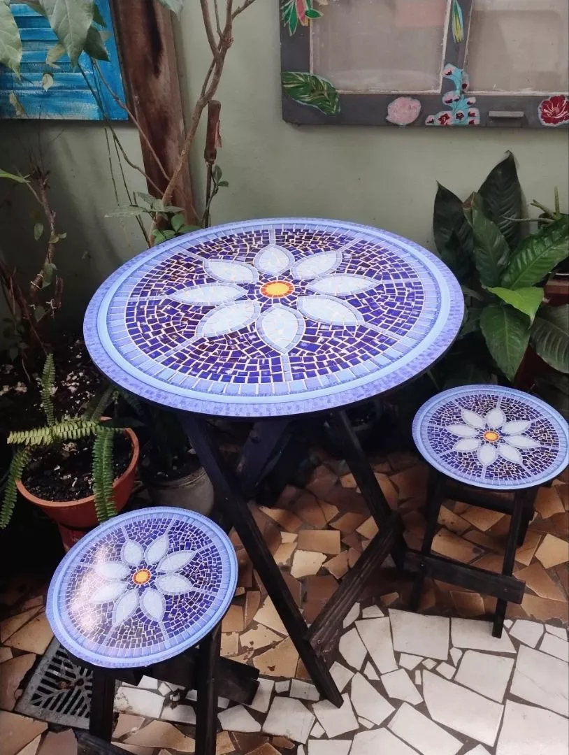 Conjunto de mesa redonda dobrável com 2 bancos em madeira escura, com tampo decorado em mosaico azul e flor branca, em uma área externa com plantas.