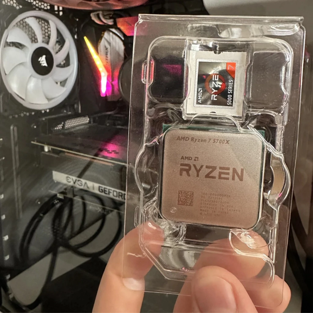 Mão segurando um processador AMD Ryzen 7 5700X dentro de sua embalagem plástica transparente, com um computador montado ao fundo.