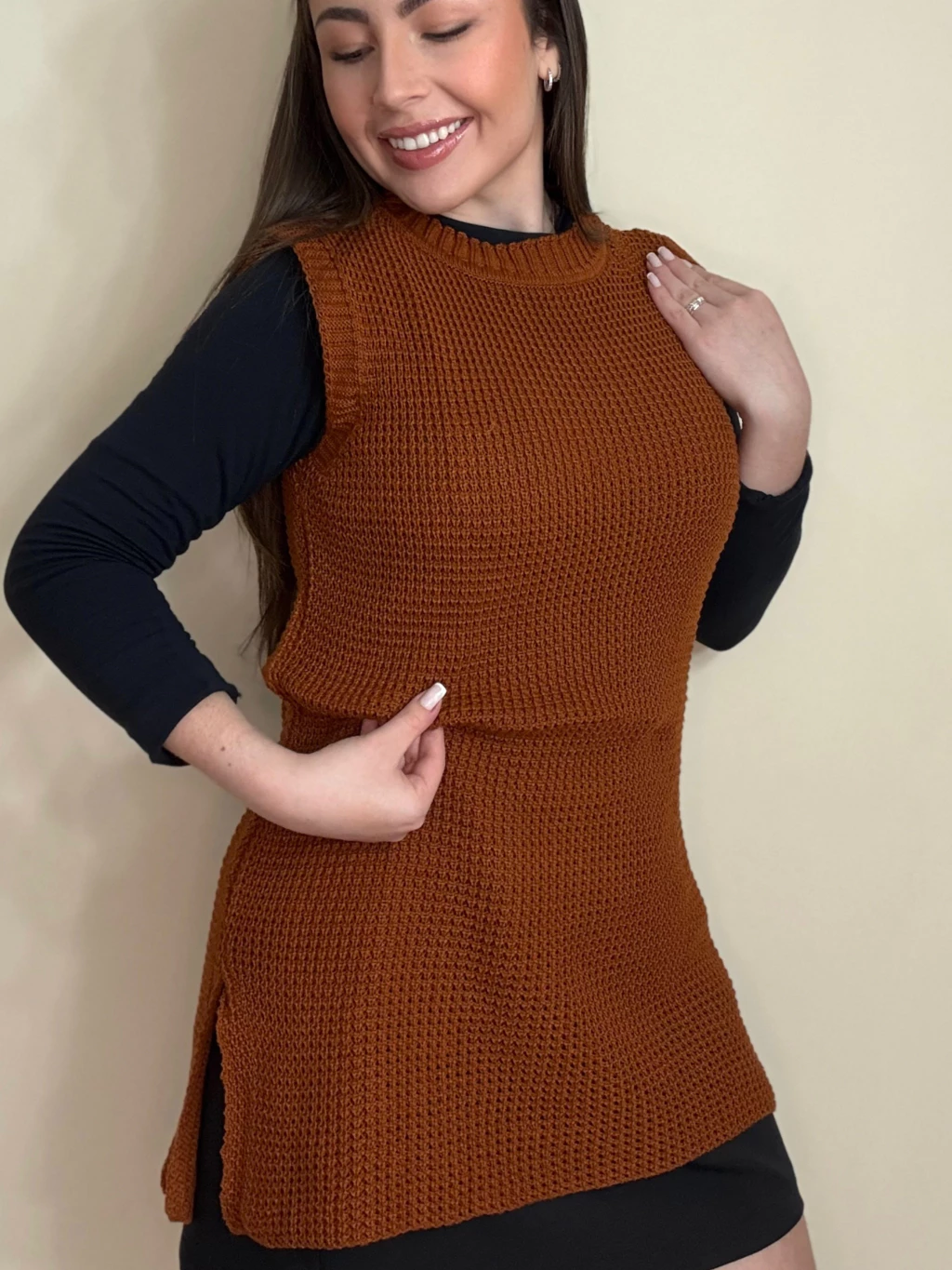 Mulher sorrindo usando colete de tricô marrom sobre blusa preta.