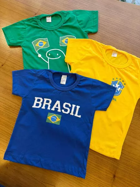 Três camisas infantis brasileiras coloridas, azul, verde e amarela, sobre uma mesa de madeira.