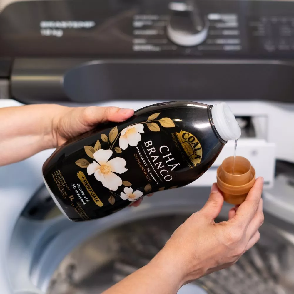 Mãos despejando amaciante Coala Laundry Chá Branco da embalagem na tampa dosadora sobre o compartimento de uma máquina de lavar