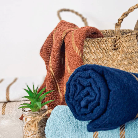 Cesto de vime com toalhas de banho nas cores azul-escuro, azul-claro e laranja-terracota, acompanhado de um pequeno vaso de planta suculenta.